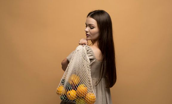 Young woman in light summer clothes with a mesh eco bag full of fruits on a green studio background. Sustainable lifestyle. Eco friendly concept. Zero waste