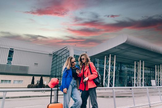 Two happy girls using smartphone checking flight or online check-in at airport together, with luggage. Air travel, summer holiday, or mobile phone application technology concept