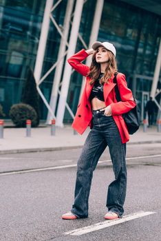 glamour woman in trendy outfit posing against the building urban background, fashion look. Outdoor fashion portrait of stylish young woman wearing black jeans, red jacket, top and a cap