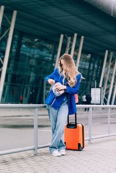 Portrait of a traveler student standing with an orange suitcase near an airport. Young fashionable woman in a blue jeans and jacket, black shirt and white sneakers. Pink baseball cap