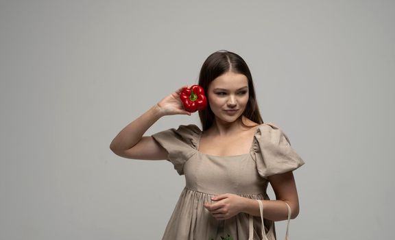 Portrait of Charming brunette woman in a beige dress with cotton mesh eco bag and a red pepper holds near a face. Zero waste shopping concept