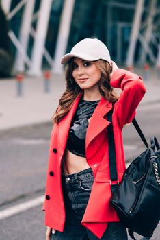 glamour woman in trendy outfit posing against the building urban background, fashion look. Outdoor fashion portrait of stylish young woman wearing black jeans, red jacket, top and a cap