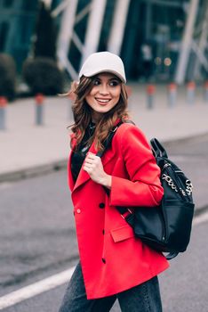 glamour woman in trendy outfit posing against the building urban background, fashion look. Outdoor fashion portrait of stylish young woman wearing black jeans, red jacket, top and a cap