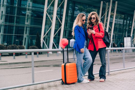 Two happy girls using smartphone checking flight or online check-in at airport together, with luggage. Air travel, summer holiday, or mobile phone application technology concept