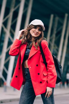 glamour woman in trendy outfit posing against the building urban background, fashion look. Outdoor fashion portrait of stylish young woman wearing black jeans, red jacket, top and a cap