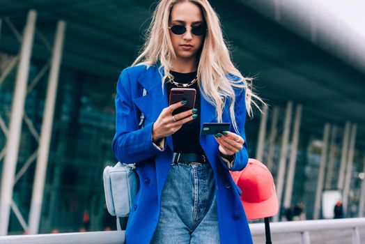 Happy fashion blonde shopper wearing a blue jacket paying on line with a credit card and a smart phone on the street.