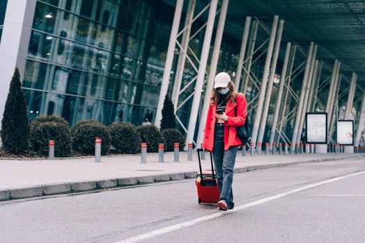 Portrait of a traveler woman in a face mask during virus epidemic walking with an orange suitcase near an airport. Young fashionable woman in a blue jeans and jacket, black shirt and white sneakers