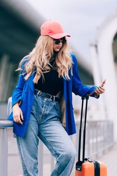 Portrait of a traveler student standing with an orange suitcase near an airport. Young fashionable woman in a blue jeans and jacket, black shirt and white sneakers. Pink baseball cap