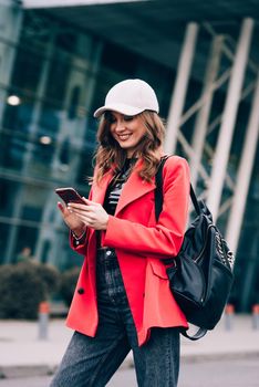 Stylish woman on the street uses a mobile phone. online shopping. use of mobile applications. beautiful woman with long dark hair in a red jacket, black top and a cap