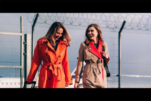 Two happy girls walking near airport, with luggage. Air travel, summer holiday. women dressed in trendy trenches orange and beige. Airport on a background