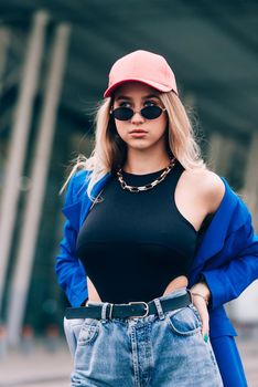 Young sexy blonde hipster woman posing on the street. Wearing blue stylish jacket, jeans and baseball hat and sunglasses. Lifestyle portrait