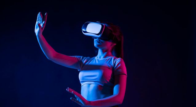 Modern architect using virtual reality glasses at workplace and touch something in virtual world. Woman working in VR goggles in neon light. Designer working in VR studio