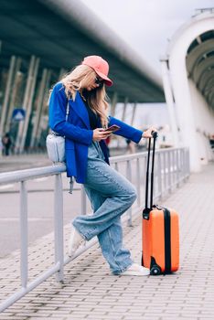 Portrait of a traveler student standing with an orange suitcase near an airport. Young fashionable woman in a blue jeans and jacket, black shirt and white sneakers. Pink baseball cap