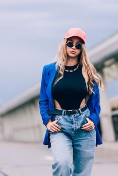 Young sexy blonde hipster woman posing on the street. Wearing blue stylish jacket, jeans and baseball hat and sunglasses. Lifestyle portrait