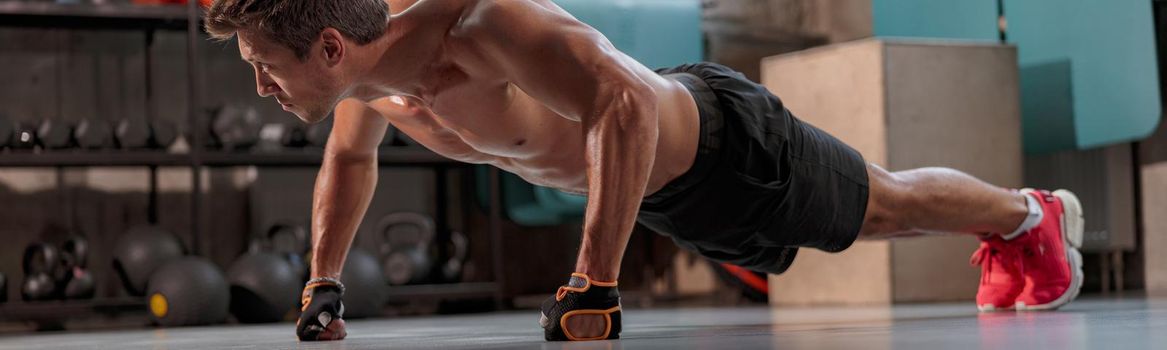 Athletic muscular man in training gloves and shorts performing sports exercise push-up from the floor in the gym