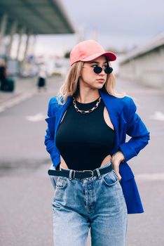 Young sexy blonde hipster woman posing on the street. Wearing blue stylish jacket, jeans and baseball hat and sunglasses. Lifestyle portrait