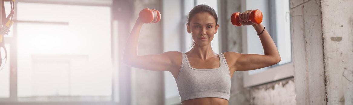 Young sporty woman holding dumbbells and performing exercise for arms, looking at camera in the fitness club