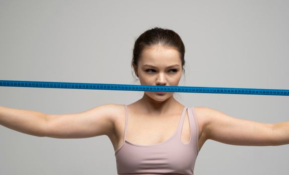 Sexy attractive brunette female in sportswear outfit holding a blue measuring tape in front of her face. Weight loss and diet concepts. Health care and healthy nutrition. Perfect slim body