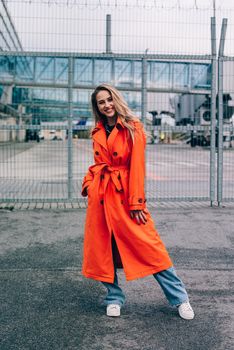 Fashionable blonde woman wearing orange coat, blue jeanse , white sneackers and neckchain posing in street of European city.