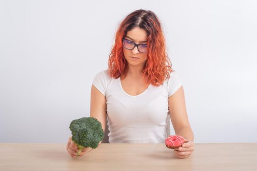 Caucasian woman chooses between vegetables and fast food. Redhead girl holding broccoli and donut on a white background