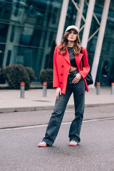 glamour woman in trendy outfit posing against the building urban background, fashion look. Outdoor fashion portrait of stylish young woman wearing black jeans, red jacket, top and a cap