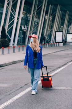 Portrait of a traveler woman in a face mask during virus epidemic walking with an orange suitcase near an airport. Young fashionable woman in a blue jeans and jacket, black shirt and white sneakers