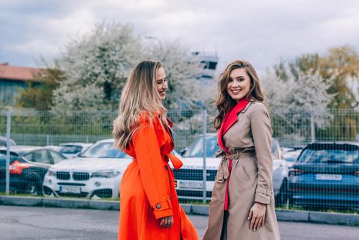 Fashionable happy smiling blonde woman wearing orange coat, blue jeanse and neckchain communicate with her girlfriend on the street. blonde and brunette happy and posing on the street