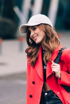 glamour woman in trendy outfit posing against the building urban background, fashion look. Outdoor fashion portrait of stylish young woman wearing black jeans, red jacket, top and a cap