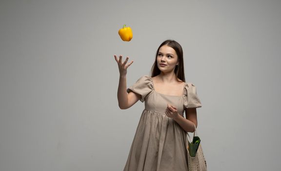 Eco friendly smiling woman in beige dress throw up a yellow pepper in a air by one hand and holding reusable cotton eco bag