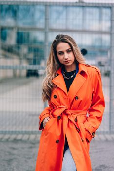 Fashionable blonde woman wearing orange coat, blue jeanse , white sneackers and neckchain posing in street of European city.