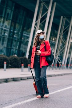 Portrait of a traveler woman in a face mask during virus epidemic walking with an orange suitcase near an airport. Young fashionable woman in a blue jeans and jacket, black shirt and white sneakers