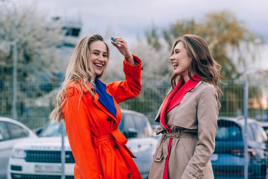 Fashionable happy smiling blonde woman wearing orange coat, blue jeanse and neckchain communicate with her girlfriend on the street. blonde and brunette happy and posing on the street