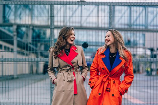 Fashionable happy smiling blonde woman wearing orange coat, blue jeanse and neckchain communicate with her girlfriend on the street. blonde and brunette happy and posing on the street