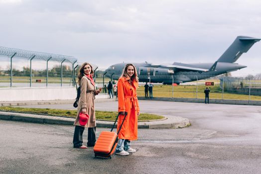 Two happy girls walking near airport, with luggage. Air travel, summer holiday. women dressed in trendy trenches orange and beige. Airplain on a background