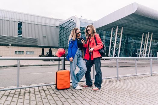 Two happy girls using smartphone checking flight or online check-in at airport together, with luggage. Air travel, summer holiday, or mobile phone application technology concept