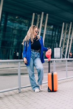 Portrait of a traveler student standing with an orange suitcase near an airport. Young fashionable woman in a blue jeans and jacket, black shirt and white sneakers. Pink baseball cap