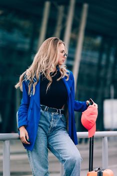 Portrait of a traveler student standing with an orange suitcase near an airport. Young fashionable woman in a blue jeans and jacket, black shirt and white sneakers. Pink baseball cap