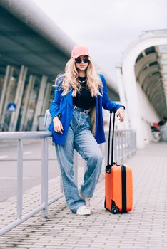 Portrait of a traveler student standing with an orange suitcase near an airport. Young fashionable woman in a blue jeans and jacket, black shirt and white sneakers. Pink baseball cap