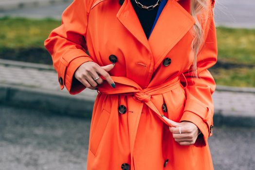 Fashionable blonde woman wearing orange coat, blue jeanse , white sneackers and neckchain posing in street of European city.