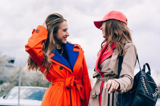 Fashionable happy smiling blonde woman wearing orange coat, blue jeanse and neckchain communicate with her girlfriend on the street. blonde and brunette happy and posing on the street