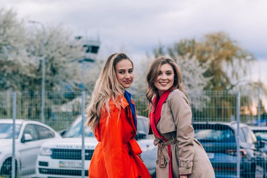 Fashionable happy smiling blonde woman wearing orange coat, blue jeanse and neckchain communicate with her girlfriend on the street. blonde and brunette happy and posing on the street
