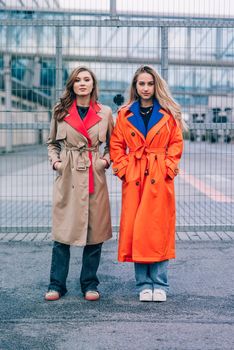 Fashionable happy smiling blonde woman wearing orange coat, blue jeanse and neckchain communicate with her girlfriend on the street. blonde and brunette happy and posing on the street
