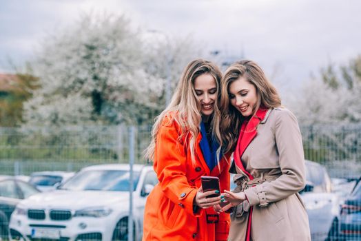 Fashionable happy smiling blonde woman wearing orange coat, blue jeanse and neckchain communicate with her girlfriend on the street. blonde and brunette happy and posing on the street