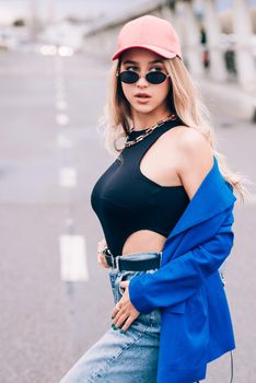 Young sexy blonde hipster woman posing on the street. Wearing blue stylish jacket, jeans and baseball hat and sunglasses. Lifestyle portrait