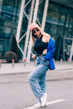 Young sexy blonde hipster woman posing on the street. Wearing blue stylish jacket, jeans and baseball hat and sunglasses. Lifestyle portrait