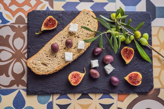 Green and black olives with loaf of fresh bread, feta cheese and young olives branch on olive wood chopping board over dark background.