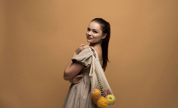 Side view of attractive happy brunette woman holding mesh grocery bag with vegetables. Concept of no plastic. Zero waste, plastic free. Eco friendly concept. Sustainable lifestyle