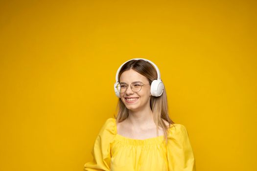 Beautiful young blonde woman in glasses with headphones listening to music and smiling and dancing, isolated on pink background