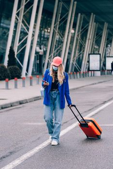 Portrait of a traveler woman in a face mask during virus epidemic walking with an orange suitcase near an airport. Young fashionable woman in a blue jeans and jacket, black shirt and white sneakers