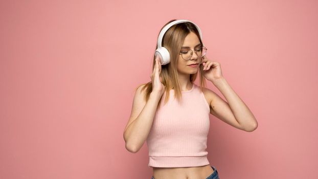 Young blonde beautiful girl in glasses in bright pink outfit enjoying a music on pink background